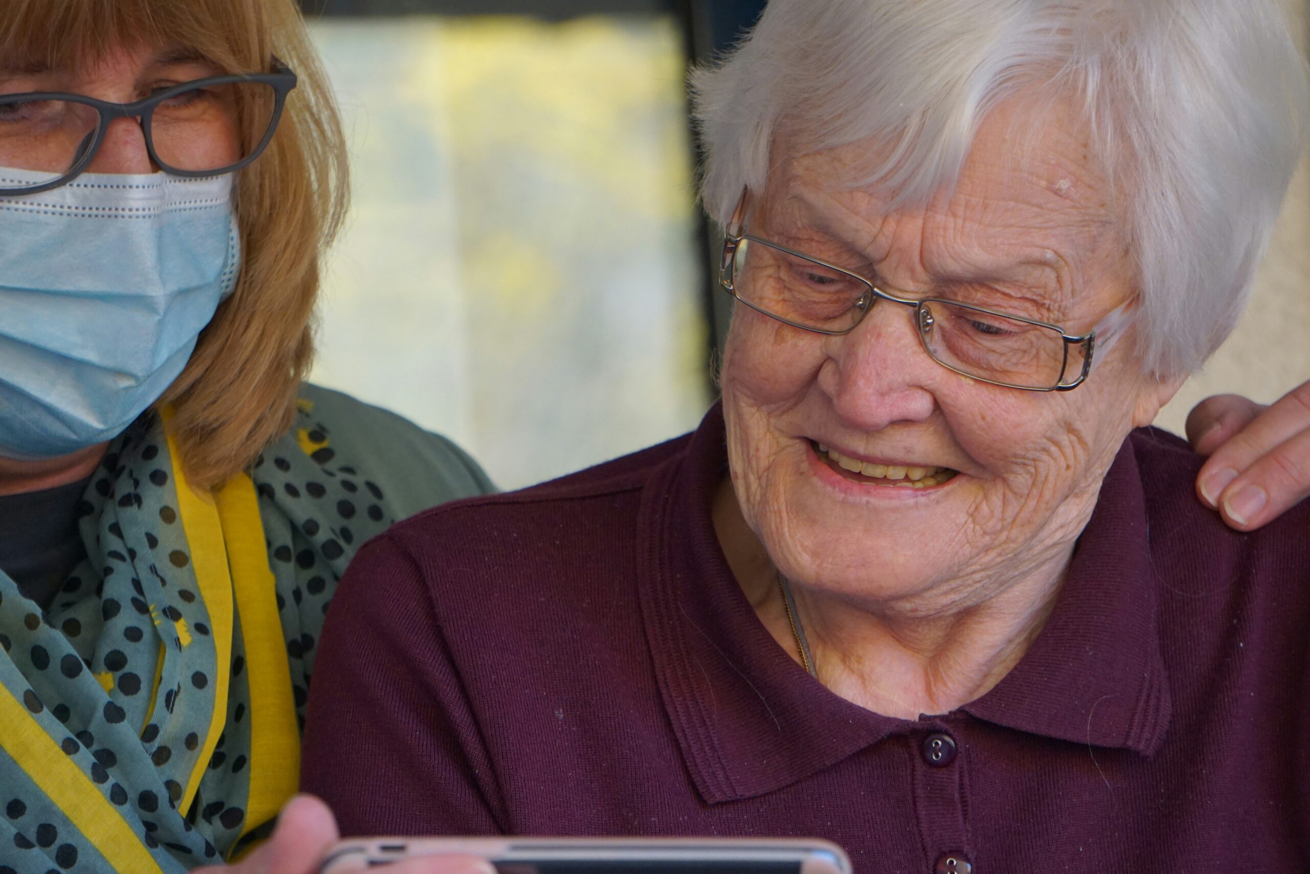 A caregiver and an elderly resident share a joyful moment as the caregiver shows something on a phone, both laughing together.