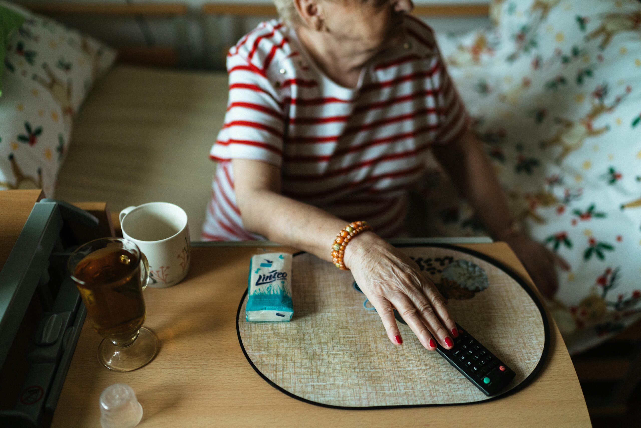 An Elderly Woman Sitting on the Bed by the Table and Holding a TV Remote Control