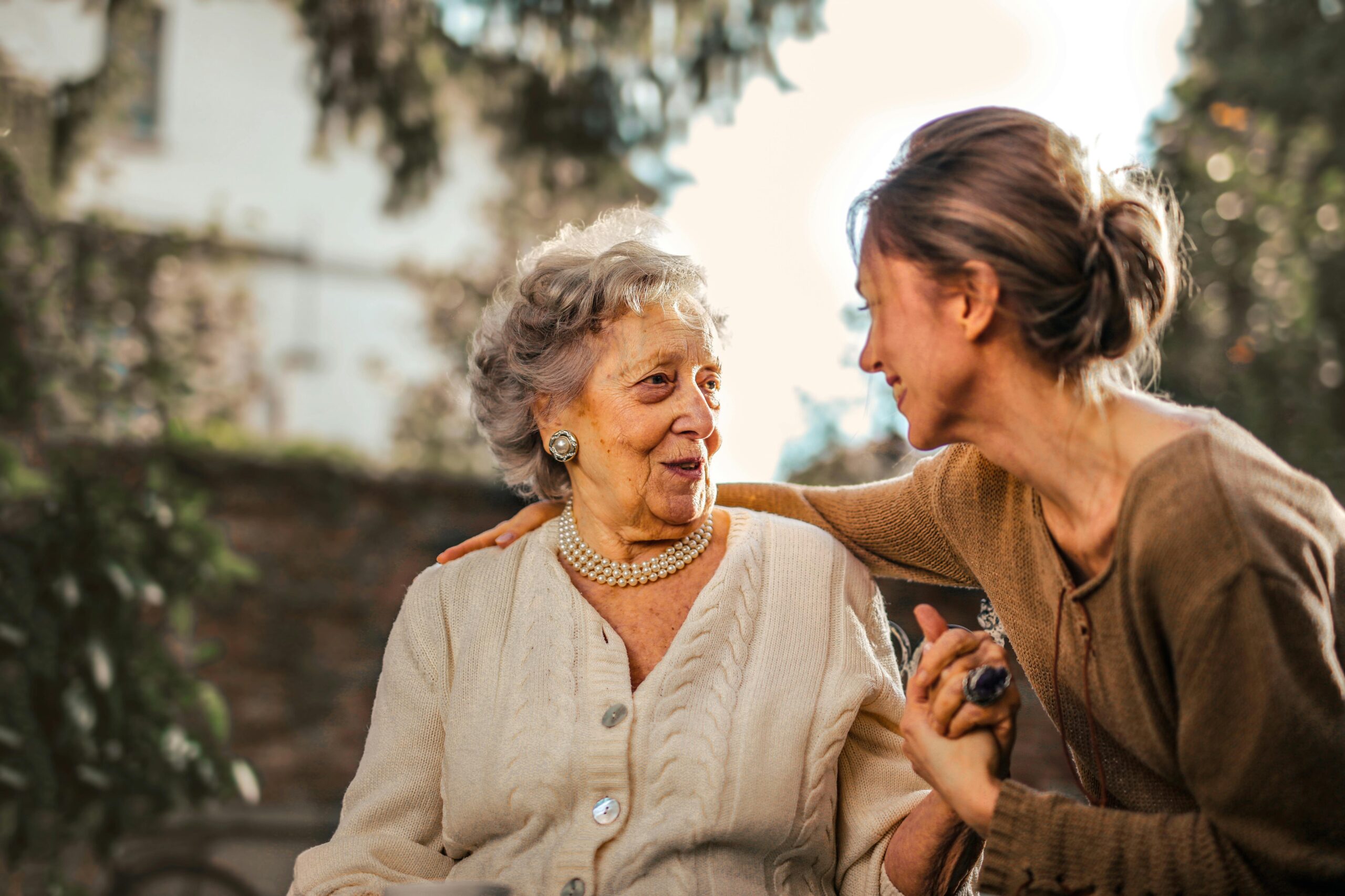 An elderly woman and a younger woman are sitting outdoors, sharing a warm moment. The elderly woman, dressed in a cream cardigan and pearl jewelry, looks delighted as she engages with the younger woman. The younger woman, wearing a brown sweater, smiles warmly while holding the elderly woman's hand and gently placing her other hand on her shoulder. The background features soft greenery and sunlight filtering through the trees, creating a serene and comforting atmosphere.