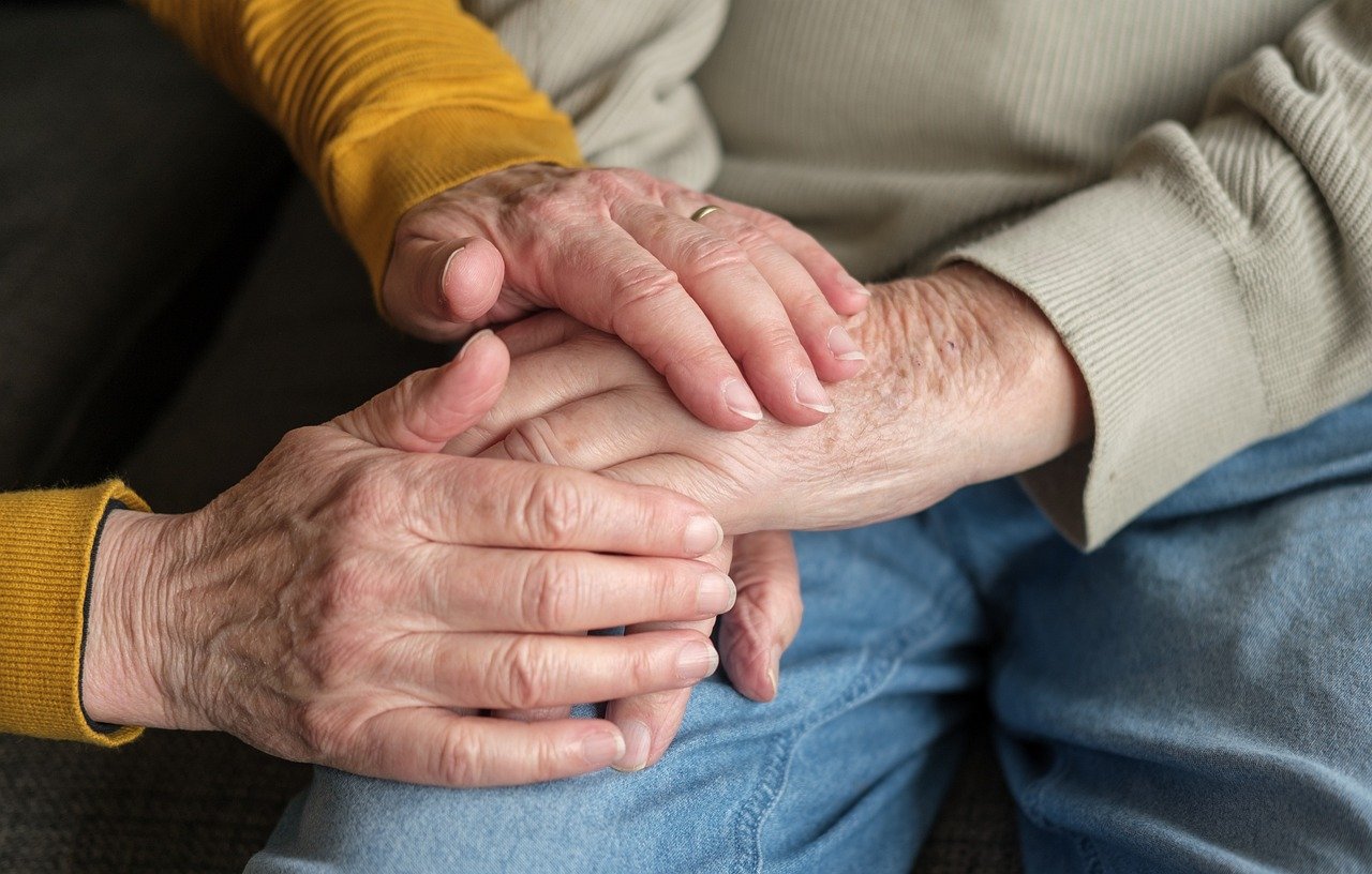 Two elderly individuals are holding hands. One person is wearing a mustard-colored sweater, and the other is dressed in a beige sweater and blue jeans. The hands convey comfort, care, and support.