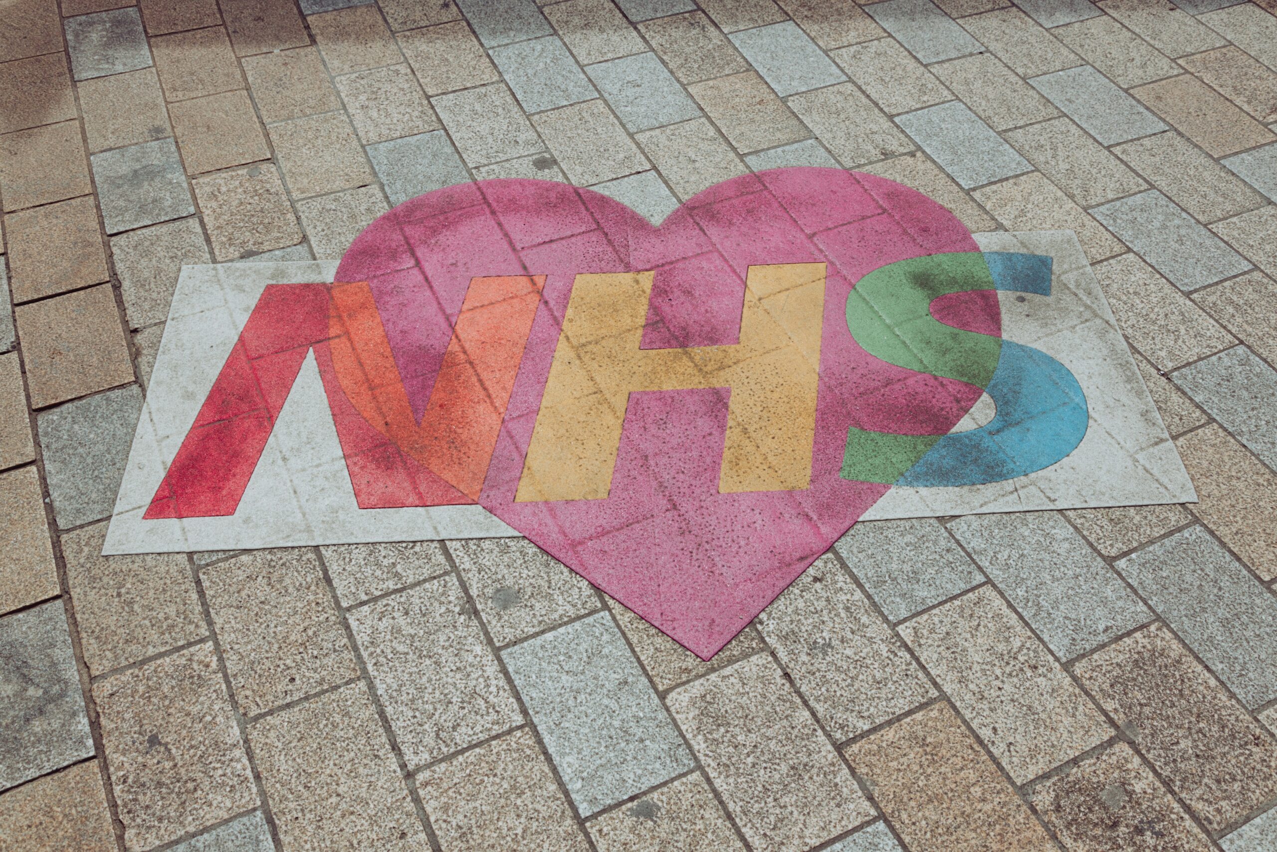 A colourful heart-shaped design on a tiled pavement with the letters "NHS" written in bold, rainbow-colored text, symbolising support for the National Health Service.