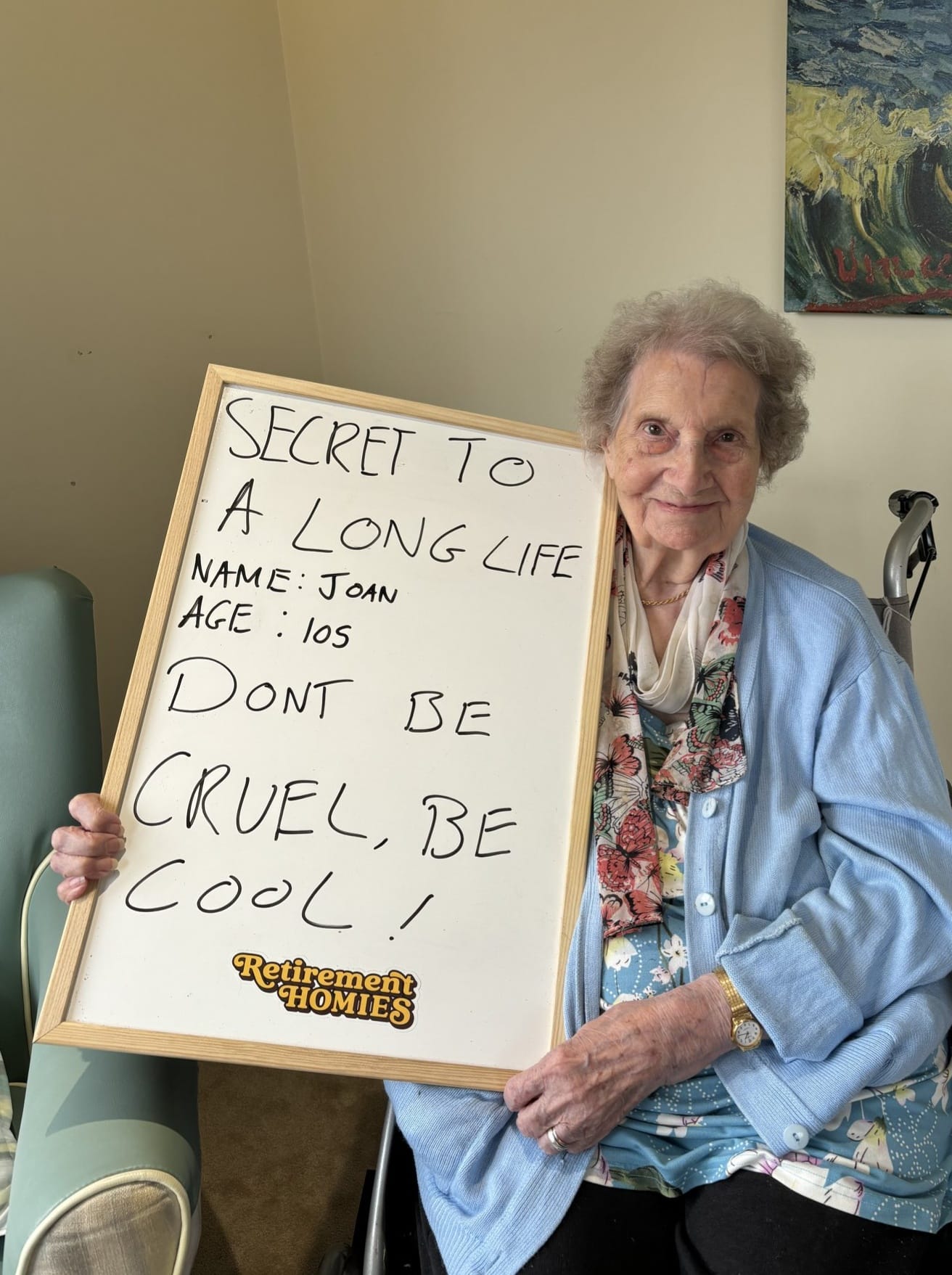 An elderly woman sitting in a Tanglewood care home is participating in the "Old Friends Club" campaign. She is holding a whiteboard that says: "SECRET TO A LONG LIFE NAME: JOAN AGE: 105 DON'T BE CRUEL, BE COOL!" She is wearing a light blue cardigan over a floral blouse and a scarf, with her hands resting on the board. A walker is visible beside her, and a colorful painting hangs on the wall in the background.