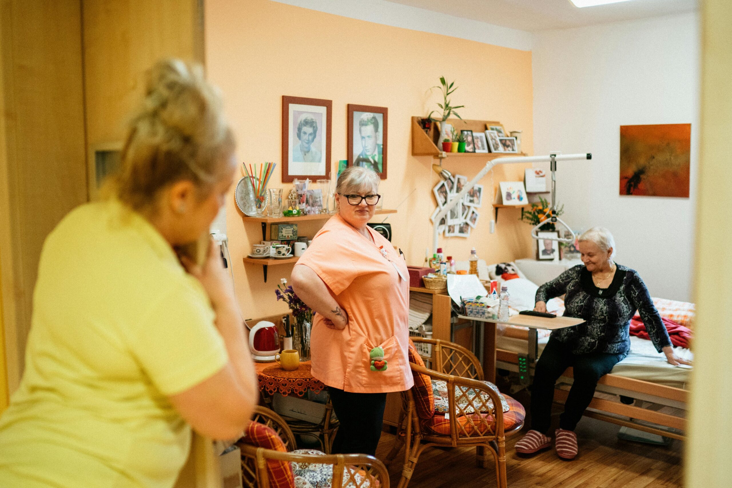 An older woman is sitting on a bed, smiling while interacting with something on a tray table in front of her. Two caregivers are present: one wearing an orange uniform stands confidently in the center of the room, and another in yellow is partially visible, standing near the doorway. The room is decorated with personal touches, including framed photos, shelves with various items, and colorful decor, creating a warm and homely atmosphere.