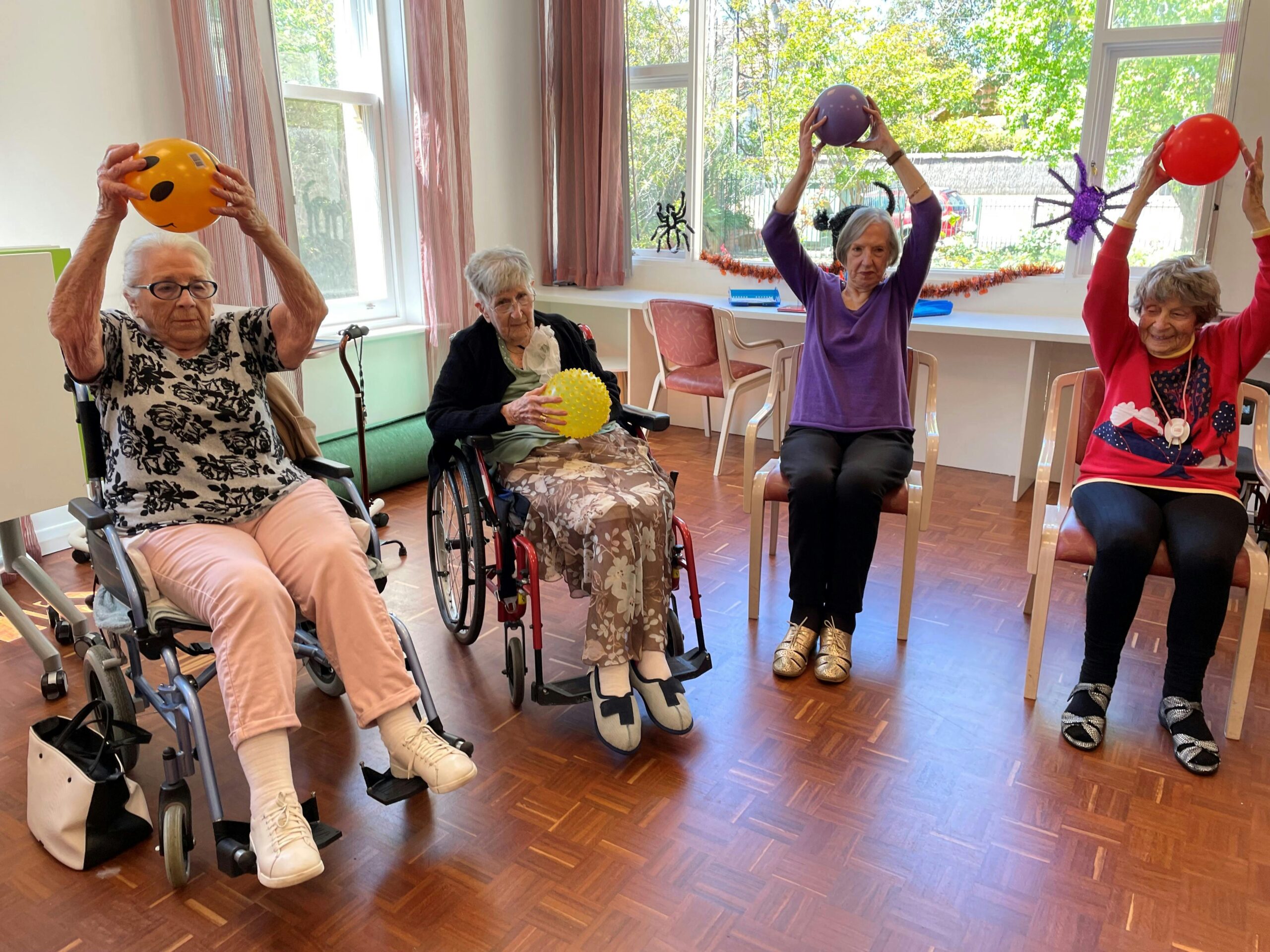 A group of elderly women is participating in a seated exercise activity in a brightly lit room. Two women in wheelchairs and two sitting on chairs are holding colorful balls above their heads, except for one who holds a yellow spiky ball at chest level. The room has wooden flooring, large windows with pink curtains, and festive Halloween decorations, including spider ornaments and garlands.