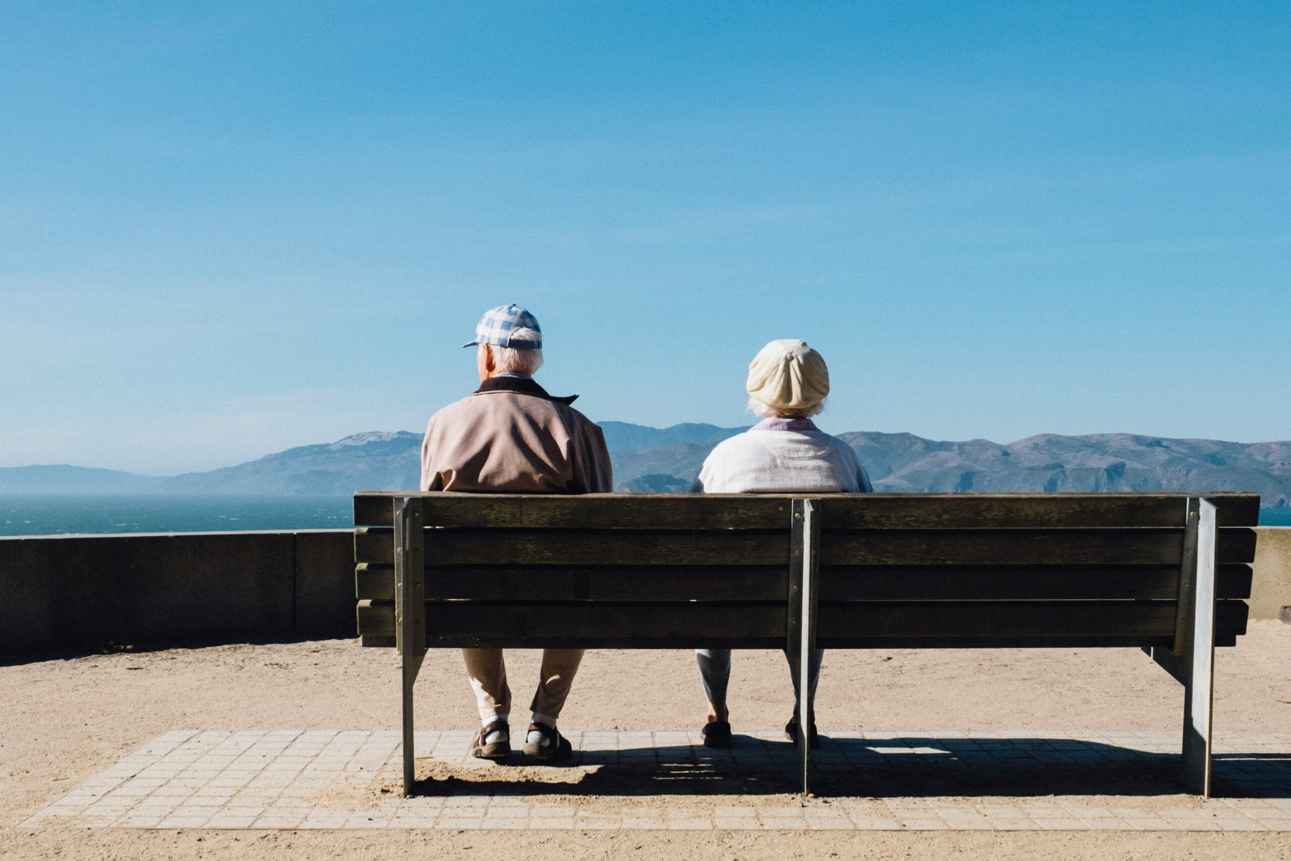 The image shows an elderly couple sitting on a wooden bench, facing away from the camera. They appear to be outdoors on a clear day, with a scenic view of mountains and water in the background. The man is wearing a light jacket and a checkered cap, while the woman is dressed in light clothing and a white hat. The setting suggests a peaceful moment of relaxation or reflection.