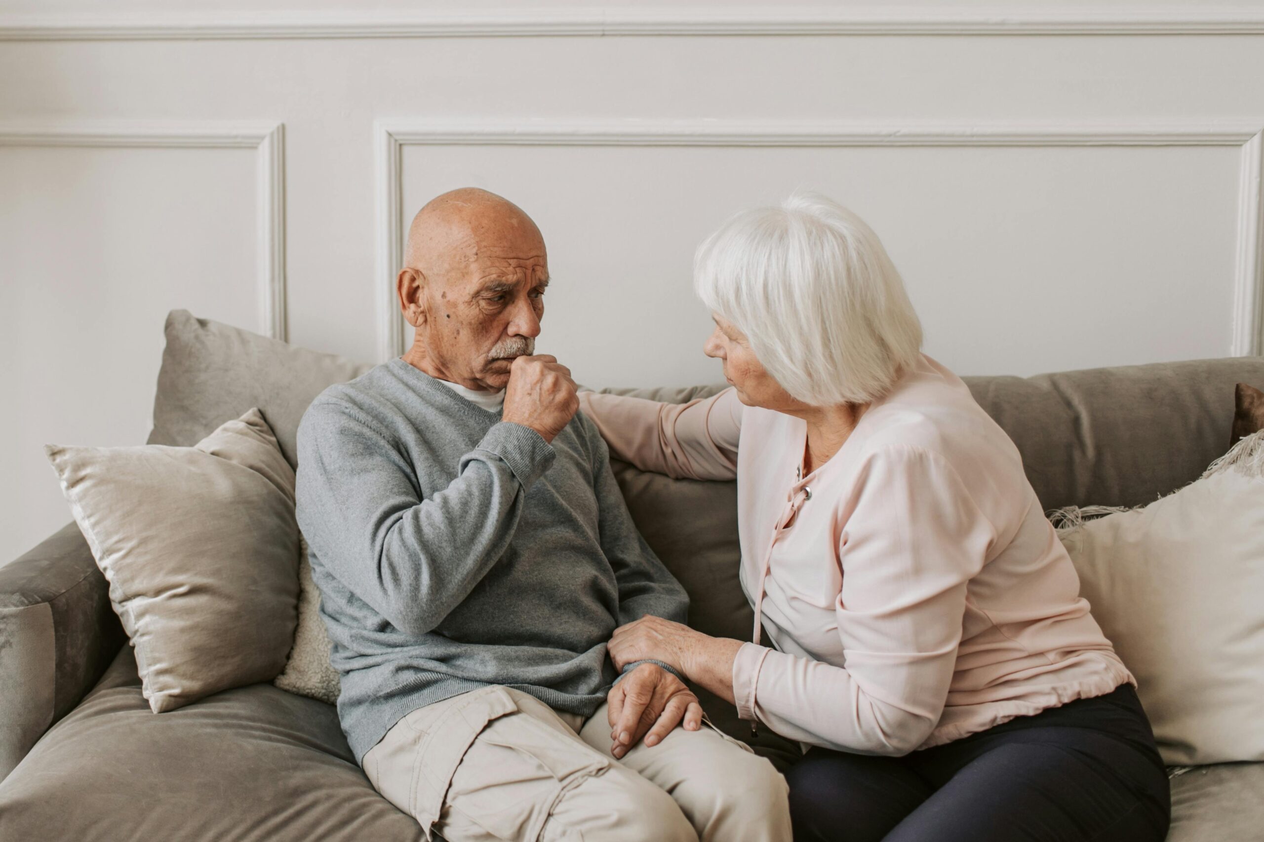 The image depicts an elderly couple sitting together on a couch in a cozy, home-like setting. The man, dressed in a light gray sweater and beige pants, appears to be receiving care or comfort from the woman, who is wearing a light pink top and dark pants. She is gently touching his hand and leaning toward him in a supportive and caring manner. The environment suggests a warm and nurturing interaction, likely emphasizing companionship, caregiving, or emotional support in later stages of life.